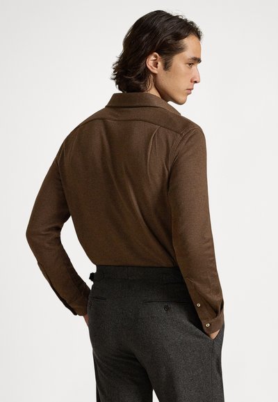 Vyras su vidutinio ilgio tamsiais plaukais, dėvintis rudą ilgomis rankovėmis marškinius ir tamsiai pilkas kelnes, stovintis šonu su rankomis kišenėse.