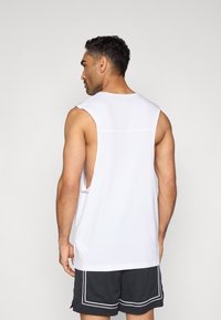 Λευκή αμάνικη μπλούζα από μαλακό ύφασμα, με βαθιά μανίκια και χαλαρή εφαρμογή, σε συνδυασμό με σκούρα σορτς και αντίθετες λευκές λεπτομέρειες.