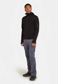 Czarna kurtka zapinana na zamek z wysokim kołnierzem, w zestawie z szarymi spodniami. Kurtka ma długie rękawy i subtelne logo na rękawie. Obuwie jest czarne.