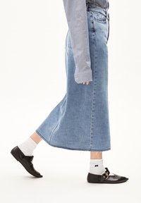 Lange spijkerrok in lichtblauw met een uitlopende vorm, ruwe zoom en zijsplitten, gedragen met zwarte schoenen en witte sokken.