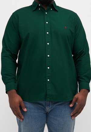 Mann trägt dunkelgrünes Hemd mit weißen Knöpfen und blaue Jeans, steht vor hellem, einfachem Hintergrund.