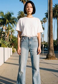 Giovane con capelli corti neri, indossa una maglietta bianca e jeans blu, in piedi su un marciapiede fiancheggiato da palme alla luce del sole.