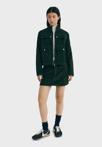 Donkergroene corduroy jas met twee voorzakken, gecombineerd met een corduroy rok. Draagt zwarte sneakers en donkere sokken.