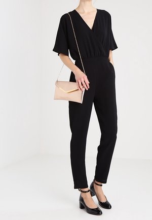 Femme en combine noire à col V portant des escarpins noirs à bride cheville et tenant une pochette beige avec une bandoulière en or.