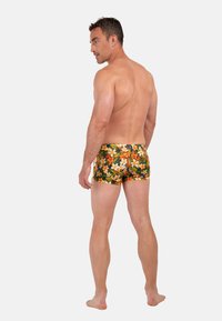 Veltrænet mand, stående barfodet med ryggen til, iført badeshorts med blomsterprint i orange og hvid på mørk baggrund.