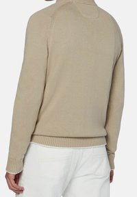 Beige neulepaita korkealla kaulaosalla, resorireunuksilla hihoissa ja helmassa. Kankaalla on teksturoitu pinta, joka korostaa yksinkertaista, minimalistista muotoilua.