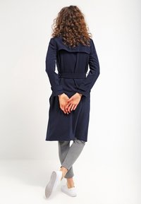 Trench coat azul marino con cinturón, mangas largas, espalda con plisado y bolsillos, combinado con leggins grises y zapatillas blancas.
