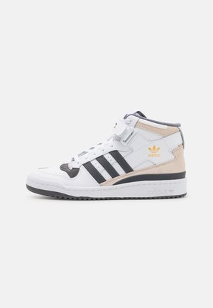 Hvid Adidas højt snøresko med sorte striber, beige ruskind detaljer og en ankelrem, vist i sideprofil mod en hvid baggrund.