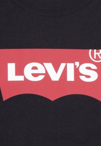 Melnas kokvilnas sporta krekls ar lielu sarkanu logotipu un baltu tekstu "Levi's", kā arī raksturīgu izliektu apakšējo malu.