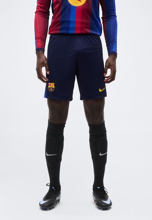 Athlète masculin portant un short de football de Barcelone, de longues chaussettes noires avec logos Nike blancs, et des crampons noirs avec des accents bleu clair.