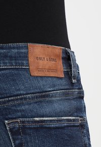 Gros plan sur une ceinture en denim foncé avec une étiquette en cuir marron portant l'inscription "ONLY & SONS" en relief, mettant en valeur des coutures détaillées et un design de poche.