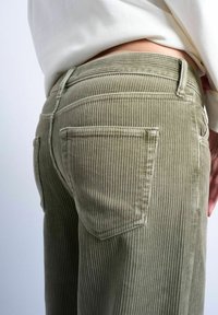 Gros plan sur une personne portant un pantalon en velours côtelé vert olive taille haute avec une poche arrière, associé à un haut blanc rentré.