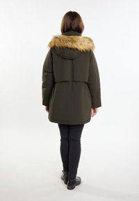 Gevoerde parka in donkergroen met een nep bontkap, voorzien van een getailleerde taille en een knielange pasvorm. Zwarte spijkerbroek en enkellaarsjes zichtbaar.