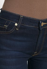 Jeans de denim azul oscuro con costuras amarillas, que cuentan con un bolsillo frontal, cierre de botón y una cintura de altura media. Textura suave con un ligero estiramiento.
