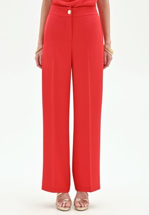 Donna che indossa pantaloni rossi a vita alta e gamba larga con bottoni frontali, abbinati a sandali dorati con cinturini e braccialetti dorati.