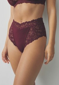 Woman wearing high-waist burgundy lace panties and matching bra, standing against a plain light-colored wall.