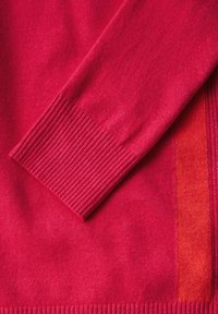Röd stickad tröja med ribbade ärmslut, som har en texturerad design och en solid färg. Inkluderar en kontrasterande panel i en något ljusare nyans.