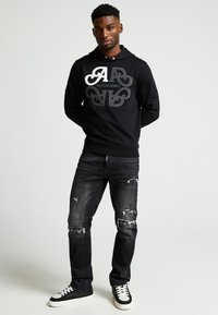 Sudadera negra con logo blanco y adornos, combinada con jeans oscuros desgastados y zapatillas negras con suelas blancas.