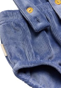 Prenda de pana azul con textura acanalada, que cuenta con dos botones amarillos y un dobladillo suave y doblado. Etiqueta visible en el lateral.