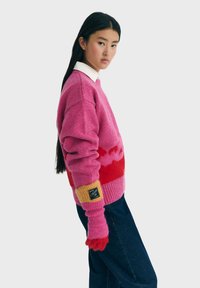 Ung kvinde med langt sort hår iført en pink og rød sweater, røde handsker, mørkeblå jeans og en hvid krave skjorte mod en lys baggrund.