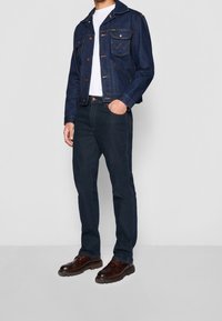 Chaqueta de mezclilla azul con dos bolsillos en el pecho, cierre de botones y cuello puntiagudo; combinada con jeans oscuros y zapatos marrones con cordones.