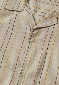 Blusa beige a maniche corte con righe verticali in rosso, nero e giallo. Presenta un colletto a revers e chiusura con bottoni. Texture del tessuto liscia.