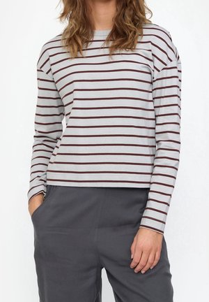 Femme portant une chemise blanche à manches longues avec des rayures horizontales marron et un pantalon gris foncé, avec une main dans la poche.