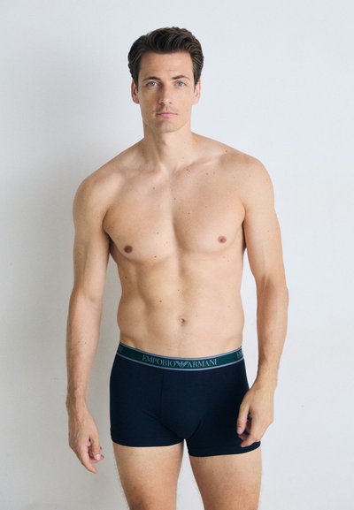 Mørkeblå boxershorts med bløt linning som har "EMPORIO ARMANI"-logo i grått. Modellen har bar overkropp og lys hudtone.