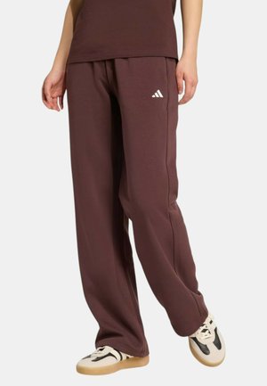 Persona che indossa pantaloni della tuta marroni larghi Adidas con logo bianco e scarpe da ginnastica bianche con dettagli neri, in piedi contro uno sfondo semplice.