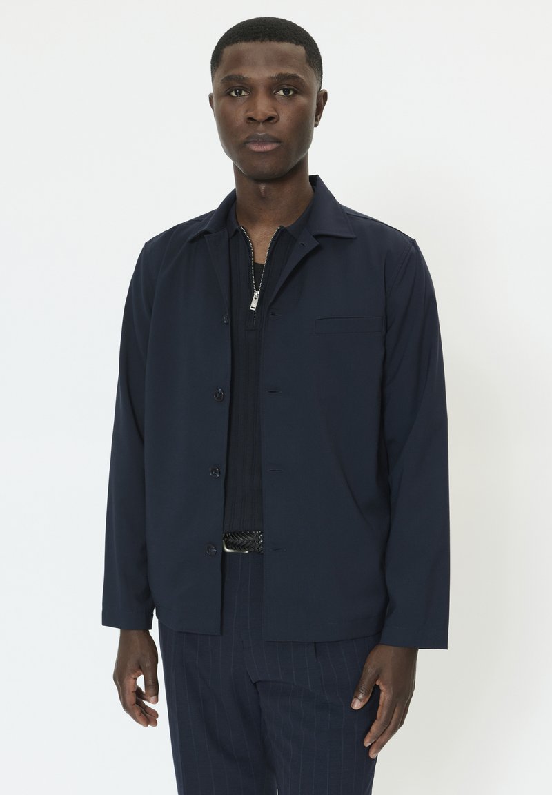 Temno modra jopica z gumbi, ovratnikom, dolgimi rokavi in prsnim žepom, nošena čez črno teksturirano majico in krojene hlače.