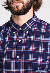 Camisa de cuadros azul marino con rayas blancas y rojas, cuello abotonado y detalle de logo en el pecho. Tela de algodón con textura suave.