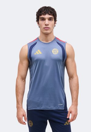 Muskularny mężczyzna stoi przodem, ubrany w niebieski bezrękawnik sportowy Adidas oraz pasujące spodnie z logo FC Bayern Monachium.
