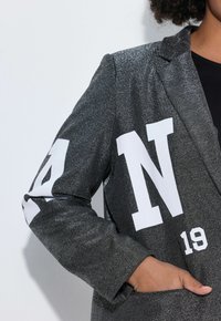 Person, die ein funkelndes dunkelgraues Blazer mit großen weißen Buchstaben und Zahlen trägt, die Hand in die vordere Tasche gesteckt, vor einem hellen Hintergrund.