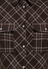 Chemise à carreaux marron foncé avec des lignes blanches et beiges, boutons-pression en nacre, deux poches poitrine à rabat et un col pointu.