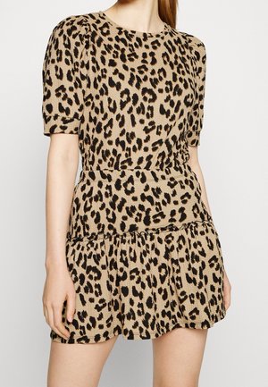 Kobieta w beżowej sukience z krótkim rękawem z czarnym i brązowym wzorem w panterkę oraz falbaną u dołu, stojąca na białym tle.