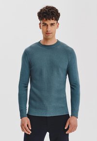 Mężczyzna w turkusowym, teksturowanym swetrze i czarnych spodniach, stojący na tle jednolitego białego tła.