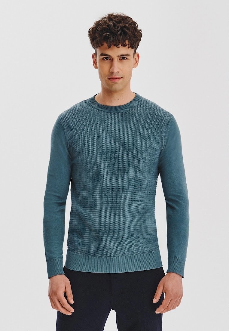 Mężczyzna w turkusowym, teksturowanym swetrze i czarnych spodniach, stojący na tle jednolitego białego tła.