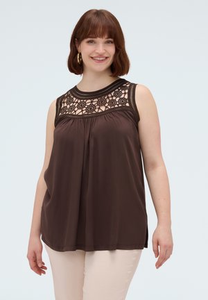 Lachende vrouw met kort bruin haar, gouden oorringen, een mouwloze donkerbruine top met bloemenkant bij de halslijn en lichte broek.
