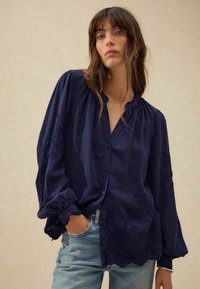 Blusa blu navy con motivi floreali ricamati sulle maniche, chiusura a bottoni frontale e scollatura con volant; realizzata in un tessuto leggero.