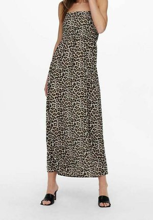 Femme portant une robe léopard bustier longue jusqu'à la cheville, avec des sandales noires à talons ouvertes aux orteils, debout devant un fond blanc uni.