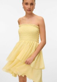 Robe jaune sans bretelles avec un corsage froncé, un ourlet à volants superposés et un tissu doux et léger, créant une silhouette fluide.