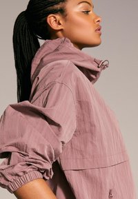 Giacca in nylon rosa chiaro con colletto alto, polsini elastici e texture sottile. Presenta una tasca frontale e un cordino regolabile nel cappuccio.