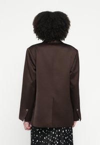 Brūns satīna žakete ar strukturētu dizainu, kas iezīmēts ar iegrieztu apkakli un vienas pogas aprocēm. Gluda tekstūra un piegriezts siluets.