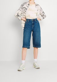 Bermuda pendek denim berwarna biru sedang, berpotongan tinggi dengan fit yang longgar. Dipadukan dengan kemeja lengan panjang tie-dye dan sepatu olahraga berwarna terang.