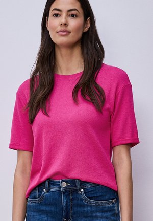 Femme aux longs cheveux noirs portant un haut en maille rose vif à manches courtes et un jean en denim bleu sur un fond clair uni.