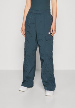 Persoon draagt donker turquoise losse cargobroek met Nike-logo op de dij en witte sneakers, staand tegen een effen achtergrond.