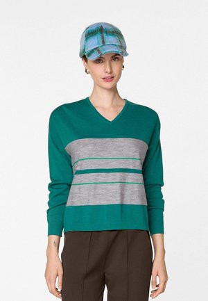 Vrouw die een teal V-hals trui met grijze en groene horizontale strepen draagt, bruine broek, blauw geruit petje en gouden oorringen, staand.