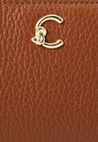 Sezione di una borsa a tracolla in pelle marrone con una superficie texturizzata e un accento del logo dorato lucido a forma della lettera "C" stilizzata.