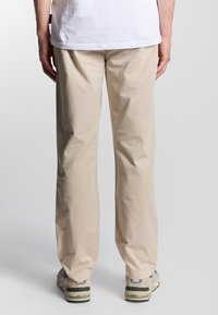 Pantaloni in cotone beige con una vestibilità rilassata, caratterizzati da una texture liscia e un design a gamba dritta, abbinati a sneakers grigie e verdi.