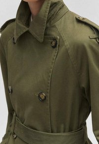 Płaszcz trencz w kolorze khaki z wysokim kołnierzem, dwurzędowym zapięciem, dużymi czarnymi guzikami i paskiem w talii. Gładka faktura tkaniny.